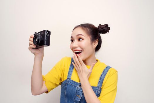 Portrait of a shocked young girl dressed in denim jumpsuit holding photo camera isolated over white background