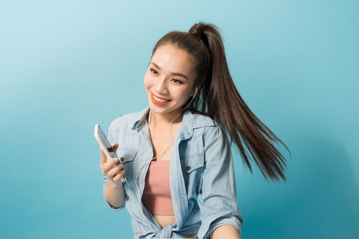 Joyous woman in casual clothing dancing and listening to music with pleasure via earphones over blue background