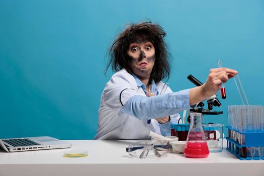 Dumb looking crazy chemist with wacky hairstyle and dirty face sitting at desk in laboratory handling glass test tubes.