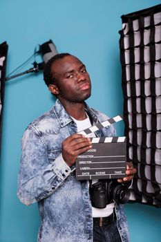 Confident producer in studio having clapperboard while standing beside professional production equipment