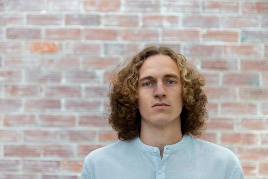 Young man looking at camera with blond curly hair in the office. Copy space. Brick wall background.