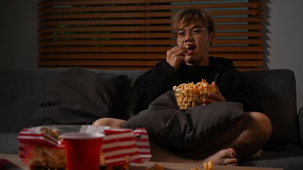 Young asian man watching TV and eating popcorn on sofa, enjoying leisure activity at home