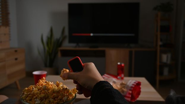 Young man with popcorn and remote control while watching movie in his living room at night