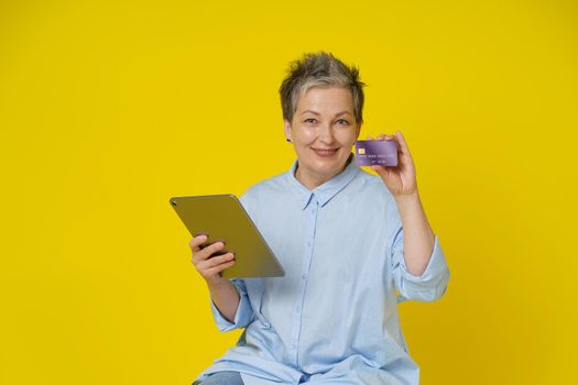 Mature woman with grey hair holding credit, debit card and tablet pc in hand making online payment or shopping online, isolated on yellow background. E-commerce, online banking concept