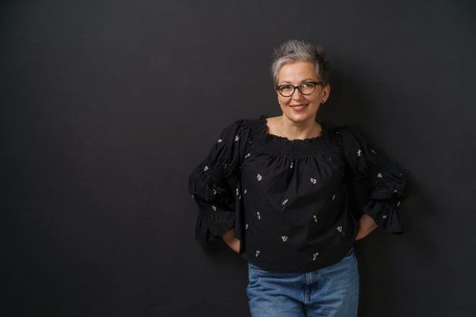 Gorgeous mature grey haired businesswoman with grey hair leaned on wall isolated on black background. Elegant woman with short hair. Image of successful mature woman. Aged beauty