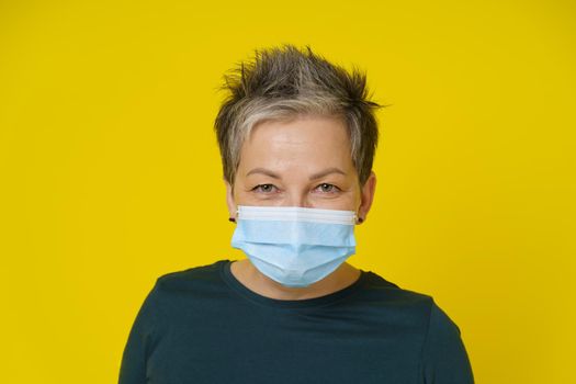 Mature woman wearing medical face mask with happy eyes smiling looking at camera wearing green blouse isolated on yellow background. Close up adult woman in medical mask