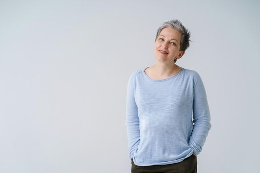 Happy dreamy mature grey haired woman 50s posing tenderly looking at camera with hands in pockets, copy space on left isolated on white background. Mature people healthcare