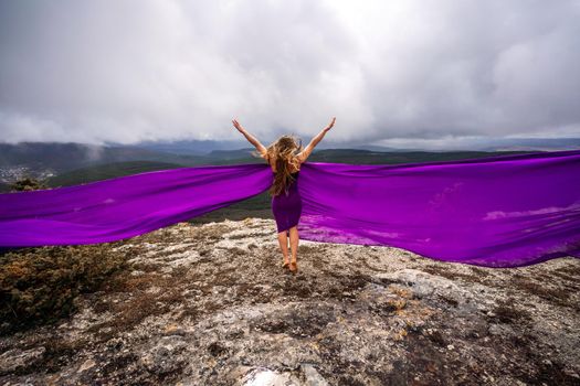 A woman with long hair is standing in a purple flowing dress with a flowing fabric. On the mountain against the background of the sky with clouds.