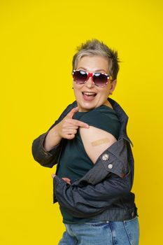 Happy mature grey hair woman showing shoulder with band aid after having shot of vaccine wearing UK flag sunglasses isolated on yellow background. Mature woman 50s vaccination and healthcare concept
