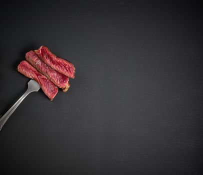 Fried pieces of beef strung on an iron large fork, black background. Meat rare