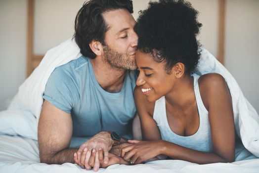 Happy, carefree and loving couple share a kiss lying in bed together in the morning. Interracial, intimate and caring husband and wife bonding and showing affection while bonding together at home