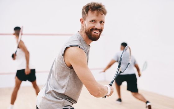 Watch me make magic with the ball. Portrait of a young man playing a game of squash with his team mates in the background.