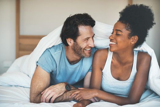 Happy, carefree and laughing couple having fun lying in bed together. Interracial husband and wife bonding and showing affection while talking. Smiling lovers enjoying time indoors being playful