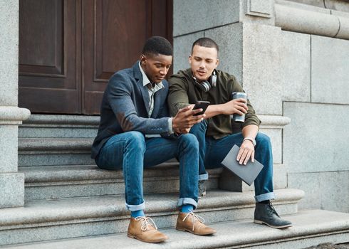 Business looks a lot more leisurely these days. two young businessmen using a smartphone together against an urban background.