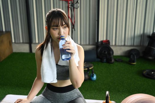 Beautiful young woman drinking water from a bottle and taking a break from exercise in fitness gym.