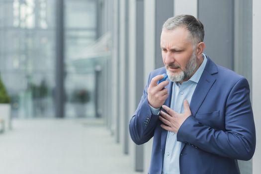 Senior male businessman sick near office, boss uses inhaler to ease breathing