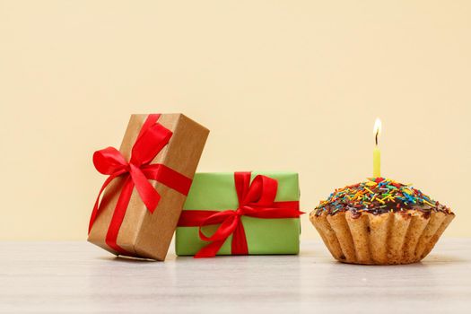 Gift boxes and tasty birthday muffin with burning festive candles on beige background.