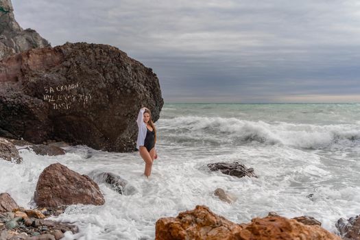 A beautiful girl in a black dress is walking on the waves, big waves with white foam. A cloudy stormy day at sea, with clouds and big waves hitting the rocks.