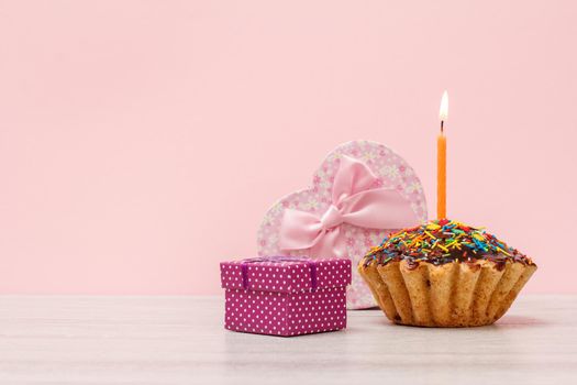 Birthday muffin with burning festive candle and gift boxes on the pink background.