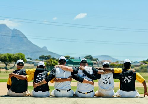 Unity always wins. Rearview shot of a team of unrecognizable baseball players embracing each other while sitting near a baseball field during the day.