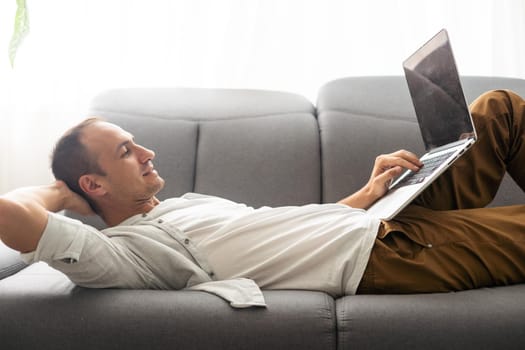 young attractive man sitting on sofa at home working on laptop online, using internet, smiling, happy mood, freelancer, free leisure time, relaxed, modern job lifestyle.
