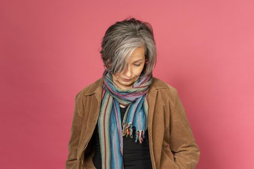 Pensive grey haired woman looks down posing in studio on pink background. Lonely concept