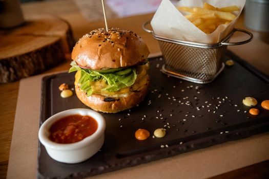 Closeup of fresh burger with basket of French fries on stone table with bowl of tomato sauce.Tasty, delicious, beef burger with fresh vegetables, fries and coleslaw