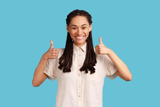 Woman with dreadlocks smiling happily, keeps thumbs up, approves something, has cheerful expression.