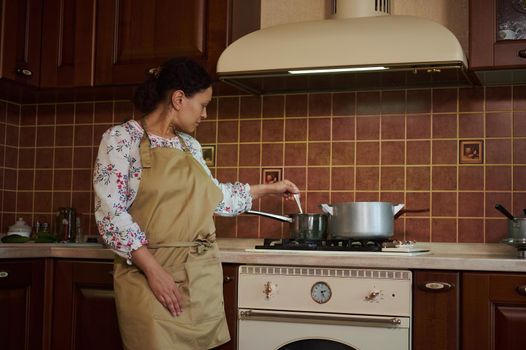 Pleasant multiethnic woman, housewife, standing at the kitchen stove and stirs pickle marinade in a saucepan