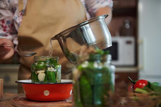 Cropped view. Housewife pouring marinade from saucepan into a jar with fresh cucumbers, while pickling in a home kitchen