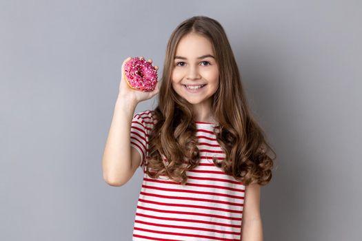 Little girl standing holding doughnut and smiling at camera, showing sweet sugary confectionary.