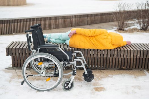 Homeless woman sleeping on a park bench next to a wheelchair.
