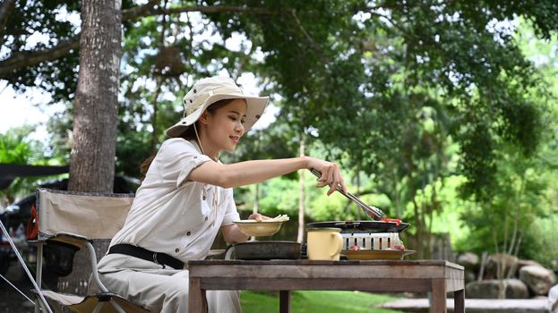 Young asian woman enjoy making barbecue during camping near the river bank. Outdoor activity or vacation concept