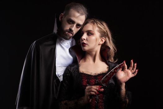 Beautiful young vampire woman with a blade covered in blood looking at her man dressed up like Dracula for halloween