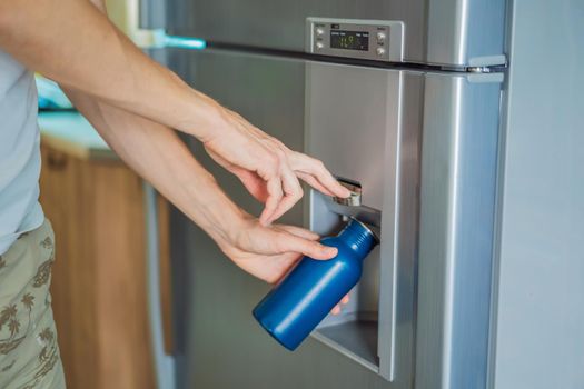 Male hand is pouring cold water and ice cubes in a metal bottle from dispenser of home fridge