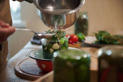Close-up of pouring boiling marinade over cucumbers in jars while pickling vegetable harvest at home kitchen for winter