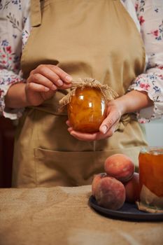 Confectioner in beige apron, holding jar of homemade jam of organic peaches, canned according traditional family recipe