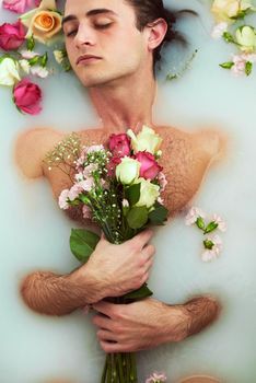 Is it even a relaxing bath without the flowers. High angle shot of a handsome young man holding a bouquet while lying in a bathtub full of milky water and flower petals at home.