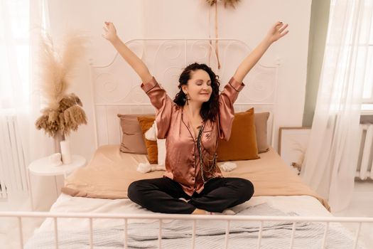 Photo of a middle aged woman in pajamas stretching her arms and smiling while sitting on the bed after sleeping or napping