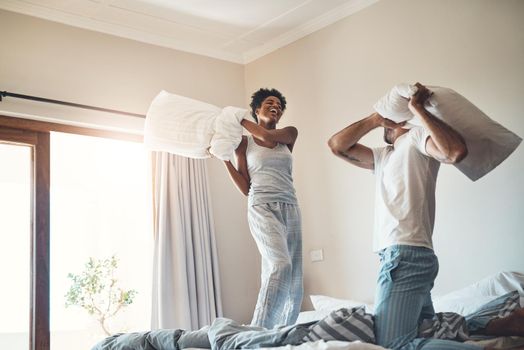 Happy, fun and playful pillow fight of a loving couple playing in their bedroom at home. Funny male and female laughing, smiling and fighting with pillows in pajamas on the bed in the morning.