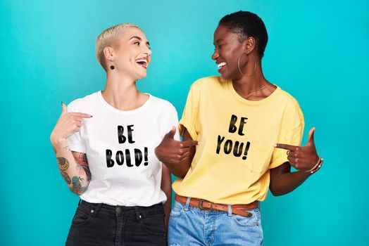 Self confidence is the biggest fashion statement. Studio shot of two confident young women pointing at their statement t shirts against a turquoise background.