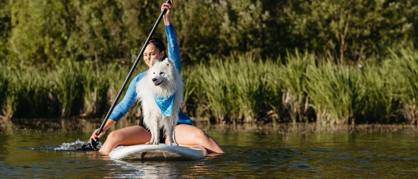 Young Woman Paddleboarding with Her Pet on the Lake, Snow-White Dog Breed Japanese Spitz Standing on the Sup Board