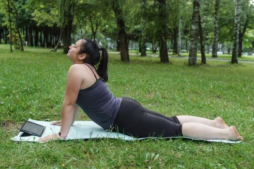 A charming brunette woman plus-size body positive practices sports in nature. Woman does yoga in the park on a sports mat.