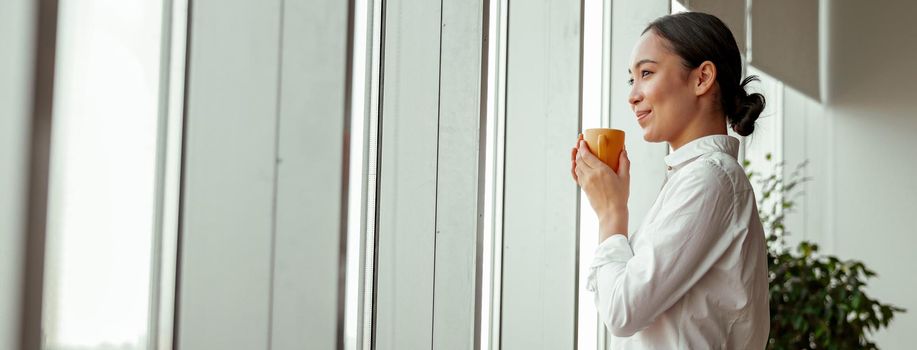 Smiling asian female worker drinking coffee standing near windows at office