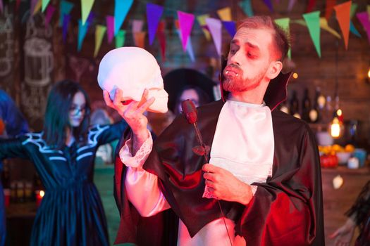Attractive young man dressed up like dracula holding a black rose looking at his human skull at halloween celebration