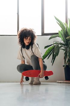 Young multiracial woman with curly hair crouching down holding a red skateboard looking down.