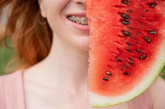Beautiful red-haired woman smiling with braces on her teeth covers half of her face with a slice of watermelon outdoors in summer.