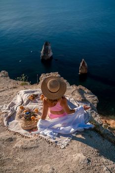 photo of a beautiful woman with long blond hair in a pink shirt and denim shorts and a hat having a picnic on a hill overlooking the sea