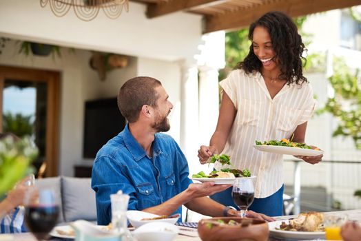 She serves every meal with a smile on her face. a beautiful young woman dishing up salad on her husbands plate while enjoying a meal with family outdoors.