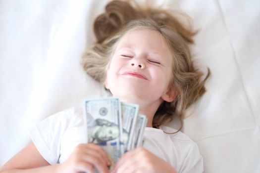 Happy girl holding cash dollars lies on bed with eyes closed